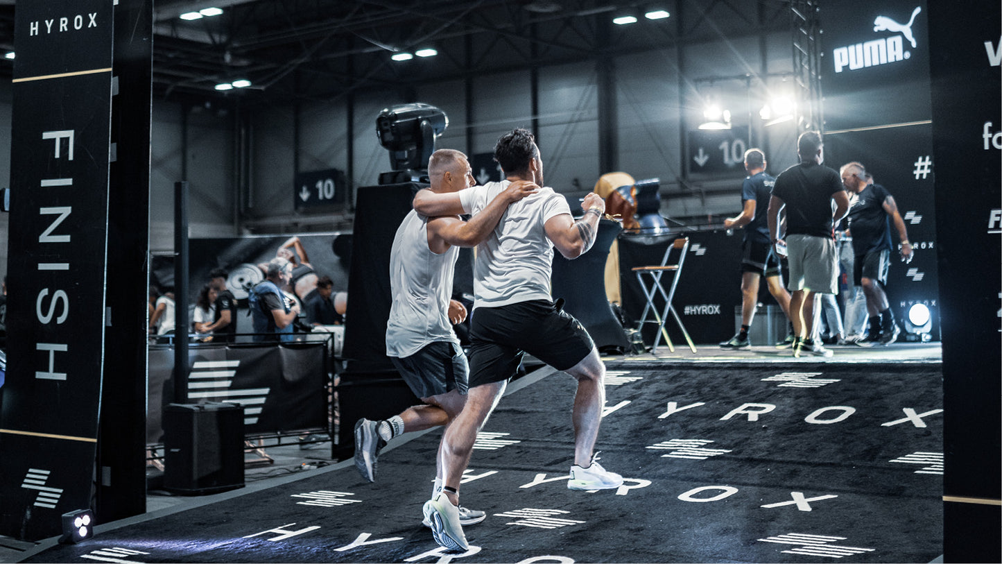 Two athletes hugging on a sports court with 'Hyrox' branding, surrounded by gym equipment and people.